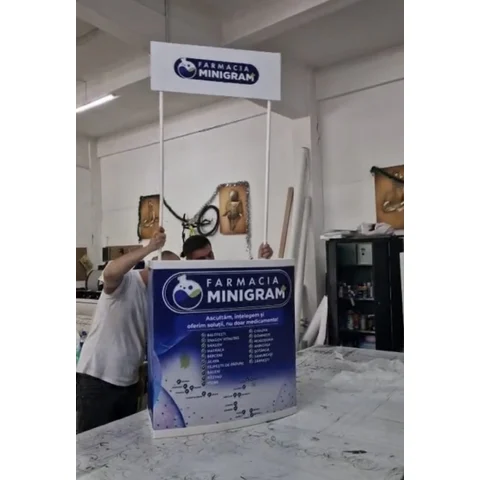 Masa expozitionala Topper Pro cu blat trapez si topper elevat, produsa de Mora Group pentru un client din reteaua de farmacii — vedere reala din atelier, panouri PVC printate full color