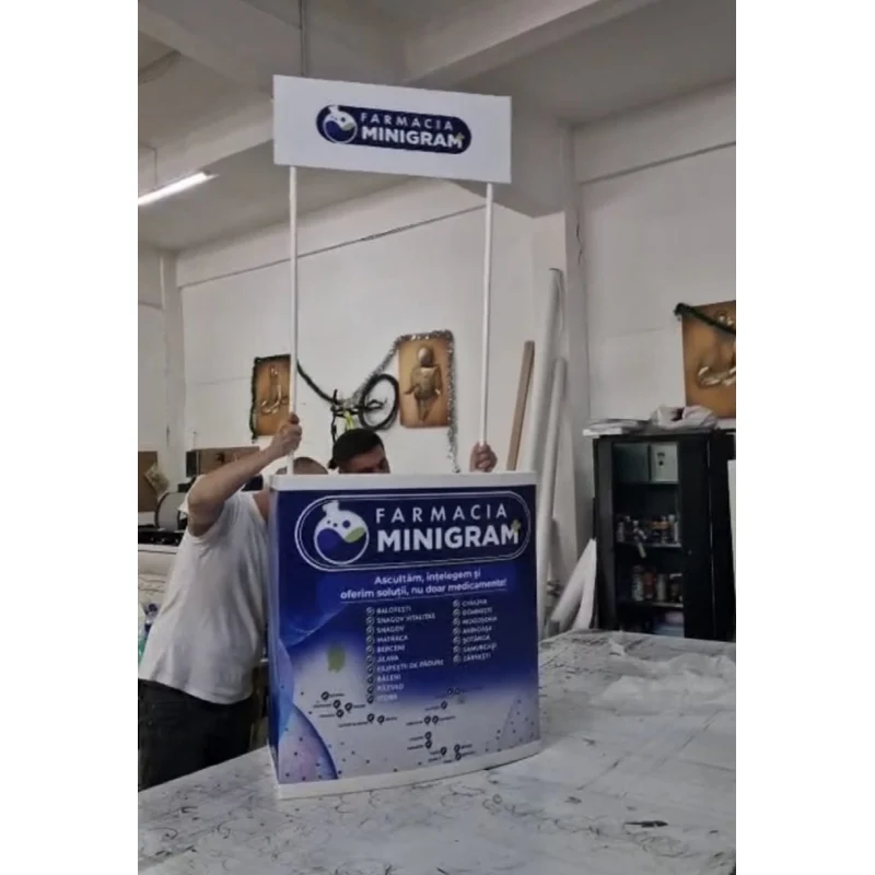 Masa expozitionala Topper Pro cu blat trapez si topper elevat, produsa de Mora Group pentru un client din reteaua de farmacii — vedere reala din atelier, panouri PVC printate full color cu logo si harta retelei