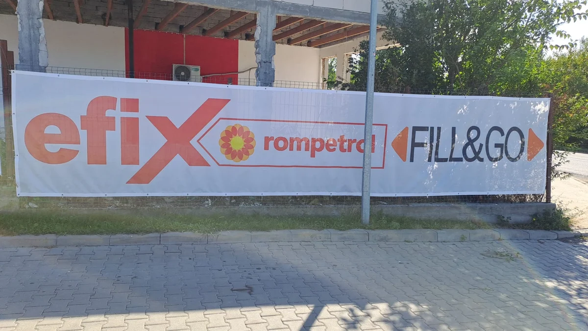 Mesh publicitar de 10 x 1,8 m produs de Mora Group, fotografiat la locatia clientului, o statie de carburant — fond alb cu logo-urile clientului si capse pe perimetru