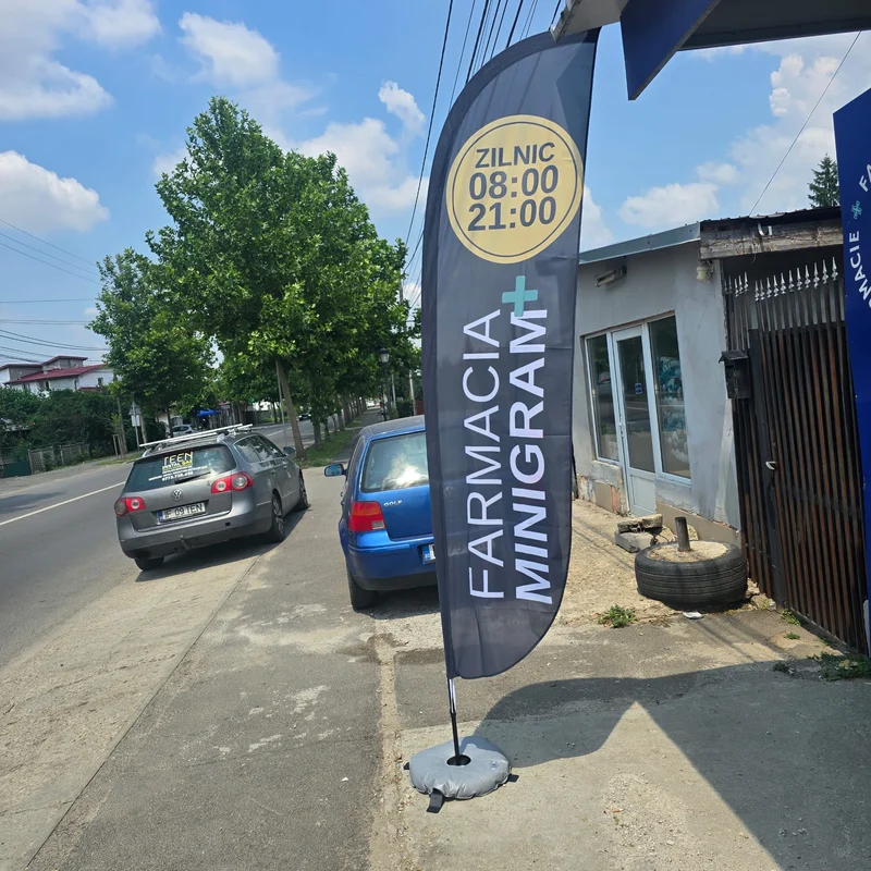 Steag publicitar tip Pana FARMACIA MINIGRAM pe suport cu baza, expus pe trotuar in fata farmaciei — sistem expozitional premium produs de Mora Group pentru promovare exterioara