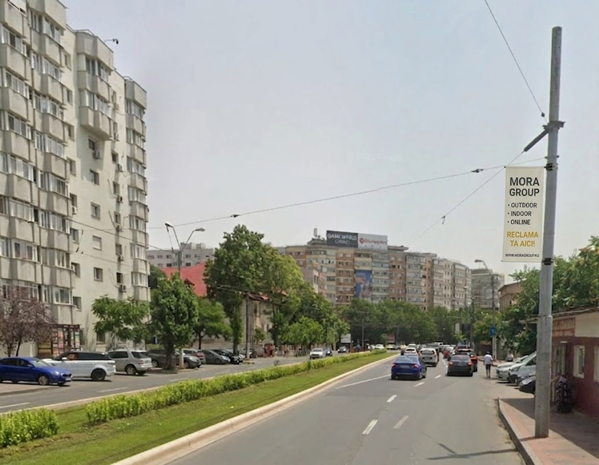 Steag publicitar Mora Group pe stalp metalic pe strada comerciala Bucuresti — zona rezidentiala cu blocuri inalte, masini parcate si trafic auto, expunere optima pentru audienta de cartier